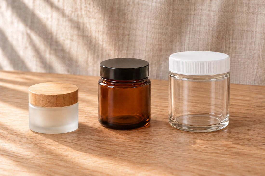 Τρία βαζάκια βάλσαμου χωρίς ετικέτα, σε διαφορετικά μεγέθη, στη σειρά πάνω σε τραπέζι από δρυ: μικρό βαζάκι από αμμοβολισμένο γυαλί με καπάκι από μπαμπού, κεχριμπαρένιο βαζάκι και μεγάλο βαζάκι από διάφανο γυαλί με λευκό καπάκι